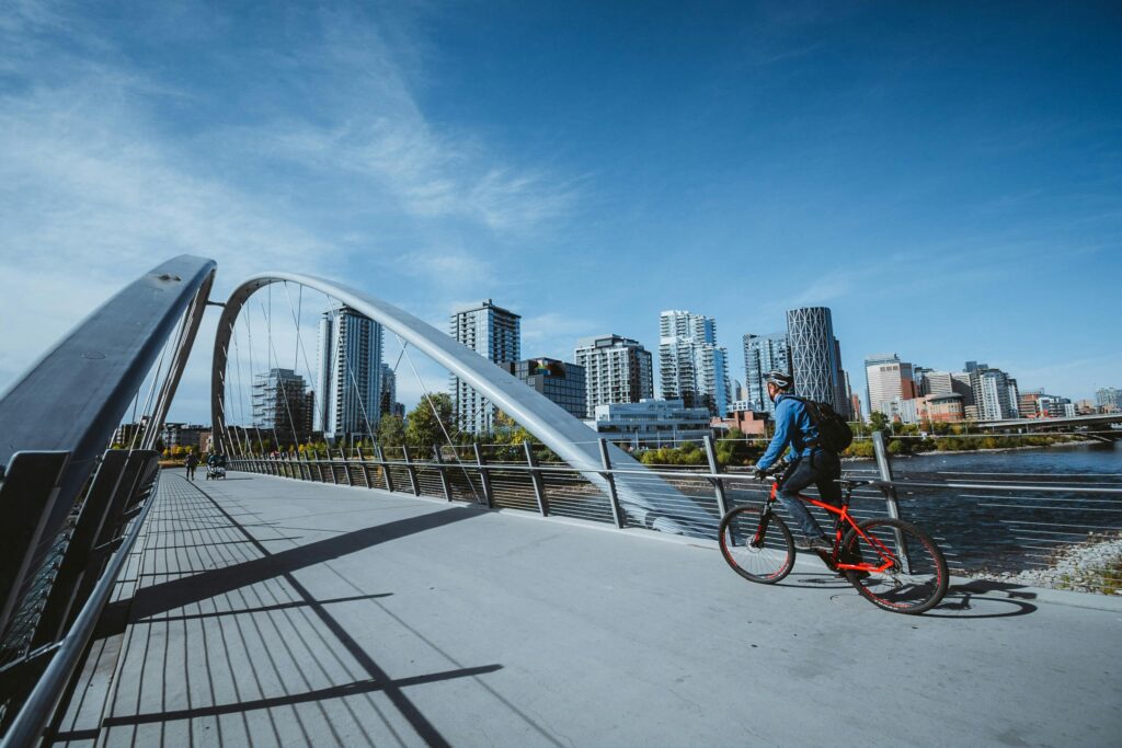 A cyclist rides across a modern bridge with a city skyline in the background, under a clear blue sky.